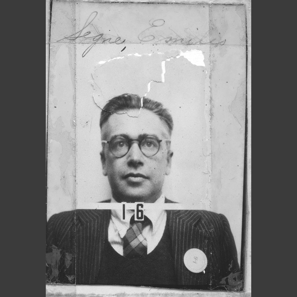 Black and white headshot of a man with round glasses wearing a striped suit, vest, and checked tie. He wears a round button on his left lapel that reads “I-6”. Written on the photo above his head reads Segre, Emilio. His badge number, I-6, also appears un