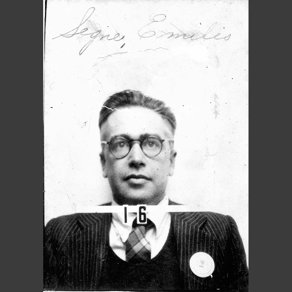 Black and white headshot of a man with round glasses wearing a striped suit, vest, and checked tie. He wears a round button on his left lapel that reads “I-6”. Written on the photo above his head reads Segre, Emilio. His badge number, I-6, also appears un