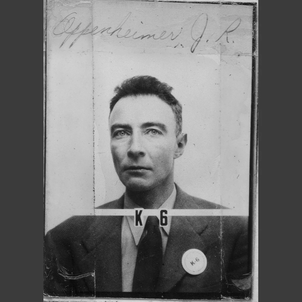 Black and white headshot of a man wearing a jacket and tie with no pattern. He wears a round button on his left lapel that reads “K-6”. Written on the photo above his head reads Oppenheimer, J.R. His badge number, K-6, also appears under his neck on the p