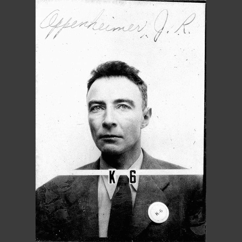 Black and white headshot of a man wearing a jacket and tie with no pattern. He wears a round button on his left lapel that reads “K-6”. Written on the photo above his head reads Oppenheimer, J.R. His badge number, K-6, also appears under his neck on the p
