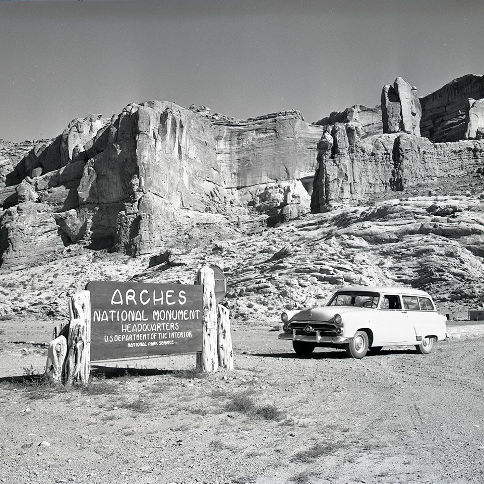 A black and white photo shows a sign reading "Arches National Monument" and a 1950s-era car sit in front of a tall rock wall in the background