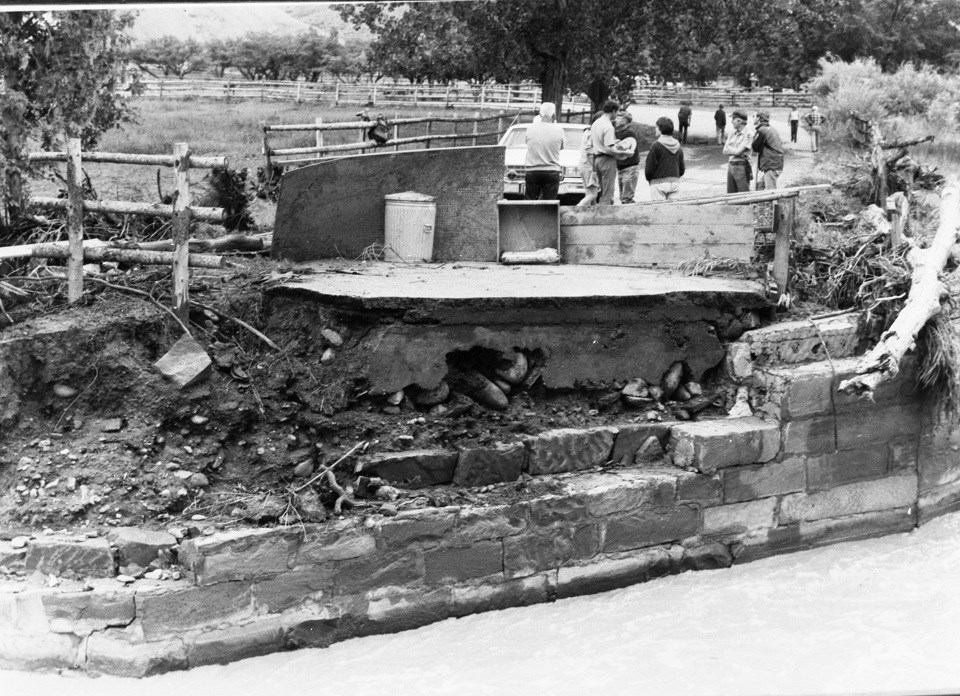 black and white photo of bridge washed out from flood, with people standing on the far side.