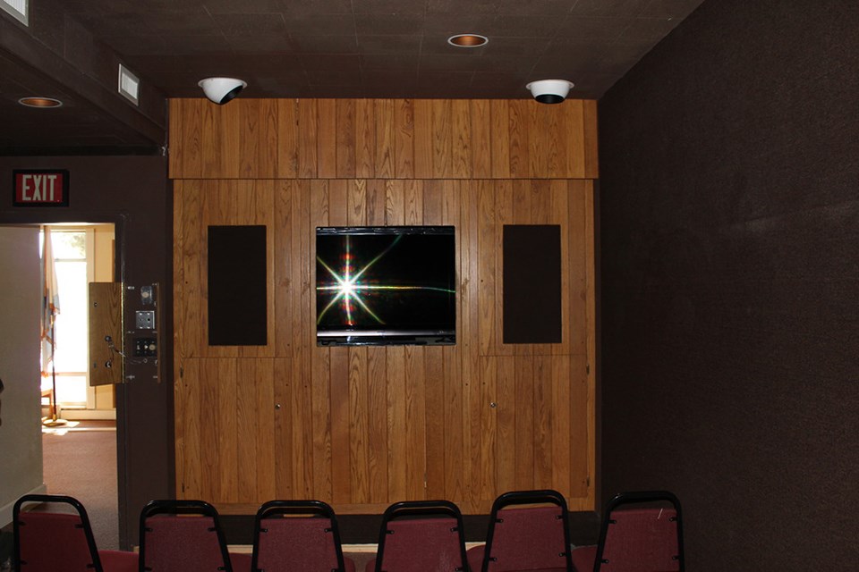 A television sits inside a wall of wood paneling.  On both sides are speakers also embedded in the wall.  Five chair backs face the camera.  To the left is an exit illuminated by daylight.