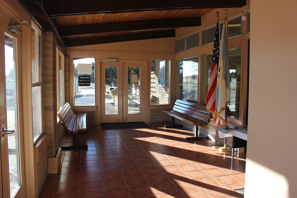 A visitor center foyer is dappled in sunlight.  Along the righ the American flag stands posted at the entry to the main building.  Ahead two benches line each side of the walkway.  To the left a brick wall of windows faces outside.