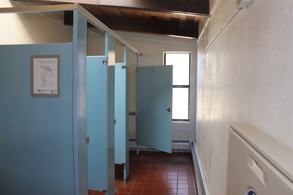 A women's restroom with three stalls along the left.  A narrow walkway leads along the stalls to a single window which lets in some light which shines faintly on the tile floor.
