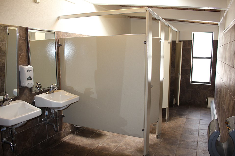 A women's restroom with three stalls along the left.  A narrow walkway leads along the stalls to a single window which lets in some light which shines faintly on the tile floor.