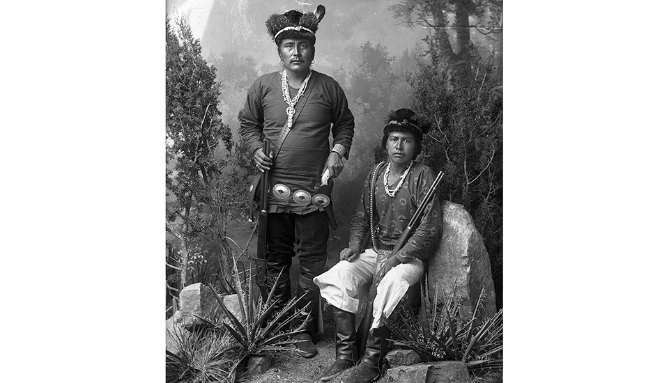 two navajo men dressed in a mixture of native and western garb holding rifles