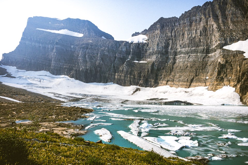 A mountainous glacier below a rocky mountain with a lake filled with icebergs in the foreground.