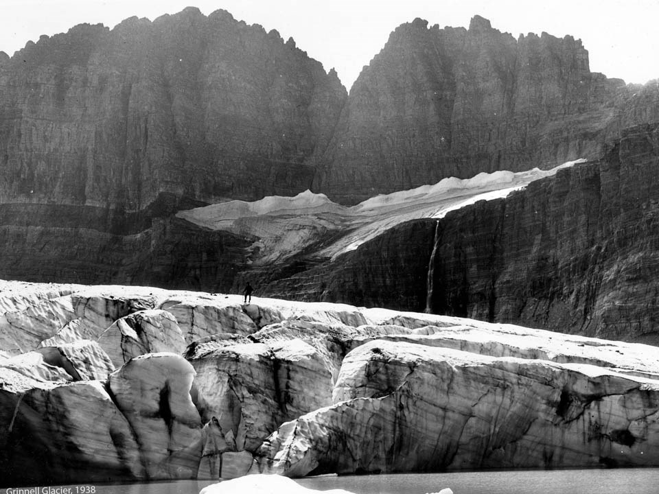 A person stands on top of a glacier in a lake with mountains in the background.