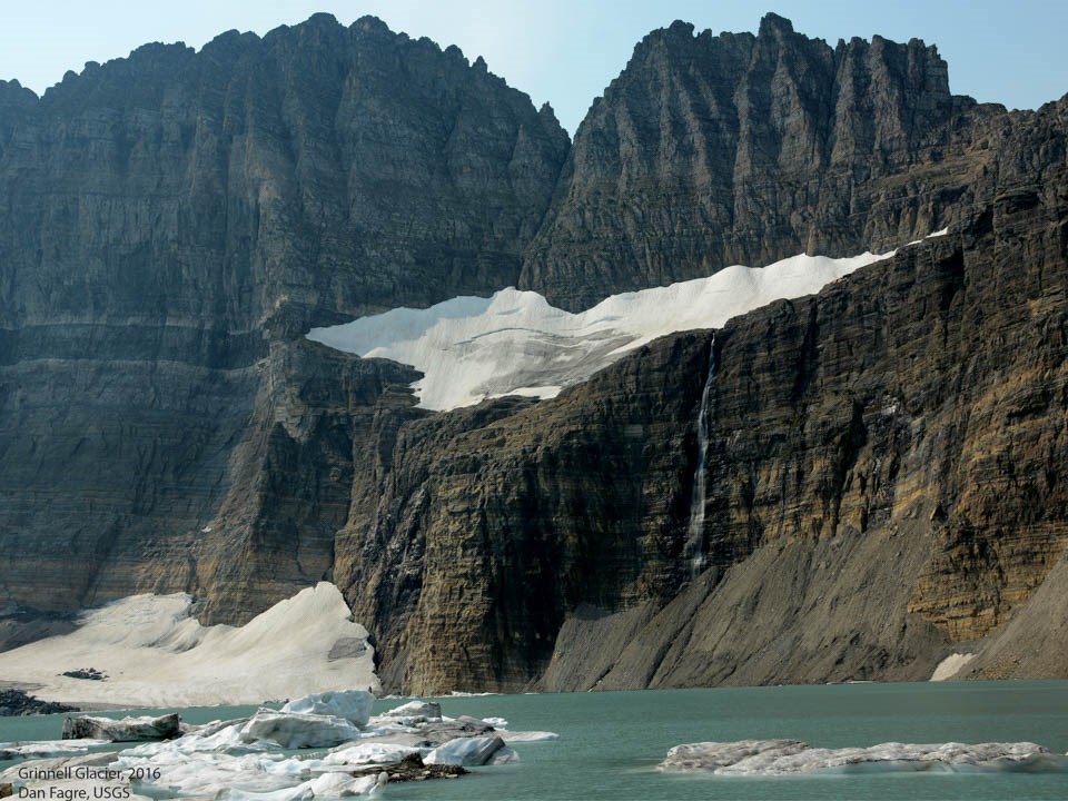 A person stands on top of a glacier in a lake with mountains in the background.