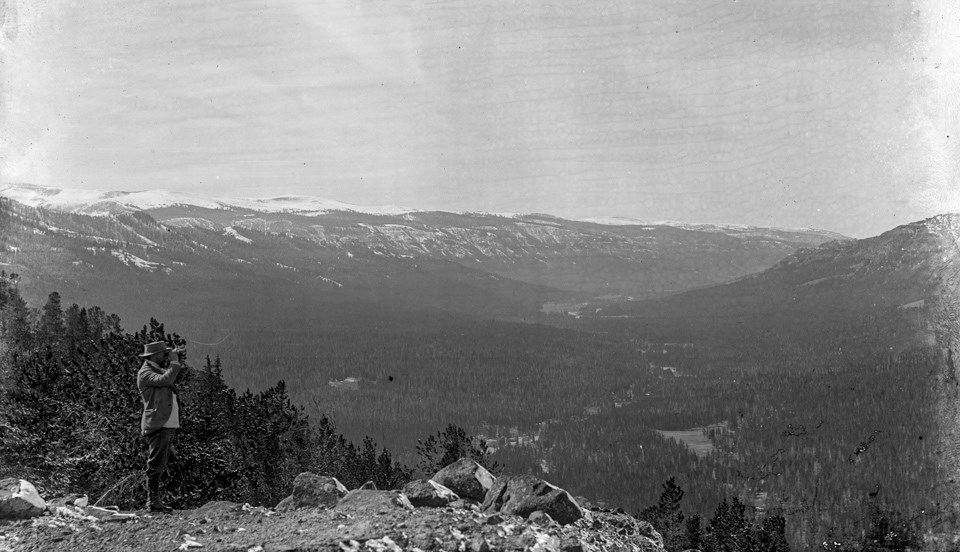 A man stands on a promontory with binoculars, looking out over a forested valley with mountains on either side and a river flowing down the center.