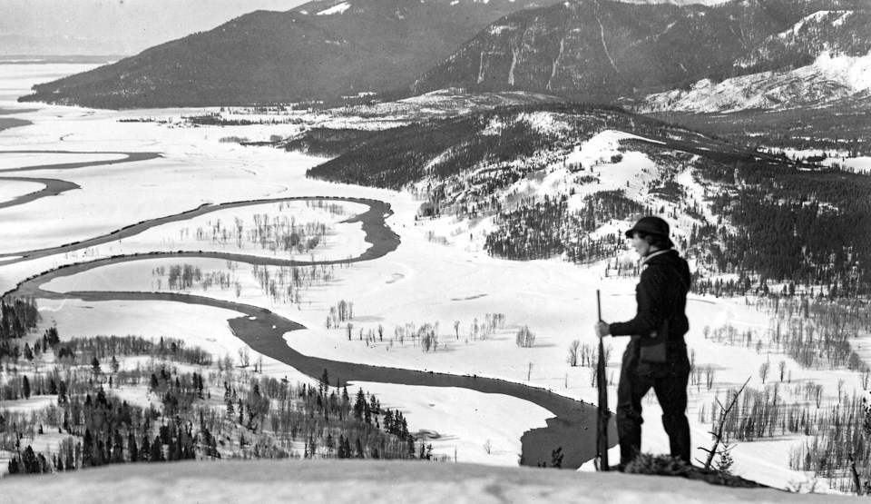 A female skier dressed in early 1900s garb stands in front of a snowy, mountainous, forested landscape above a lake.