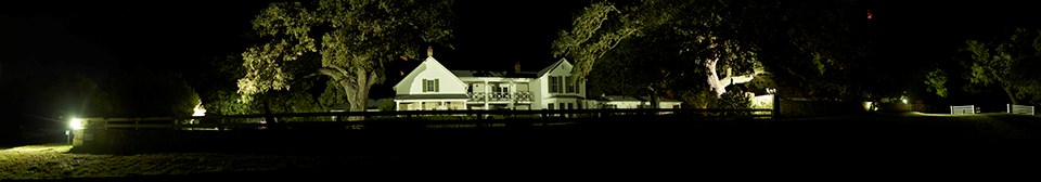 A large, two-story white house glows at night from exterior lighting.