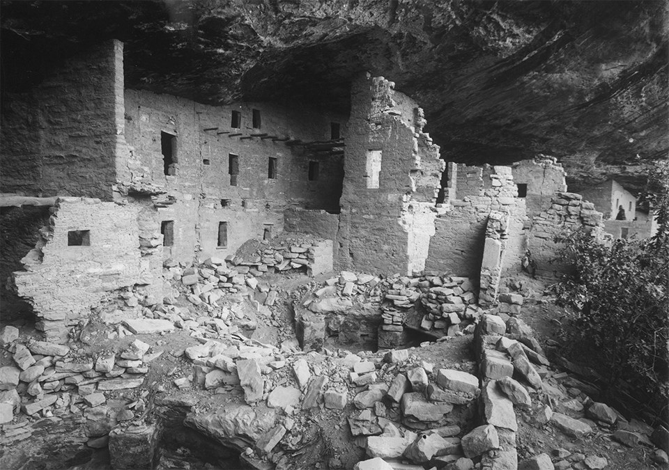 Ancient, stone-masonry village rooms under an alcove with crumbled walls and piles of rubble.