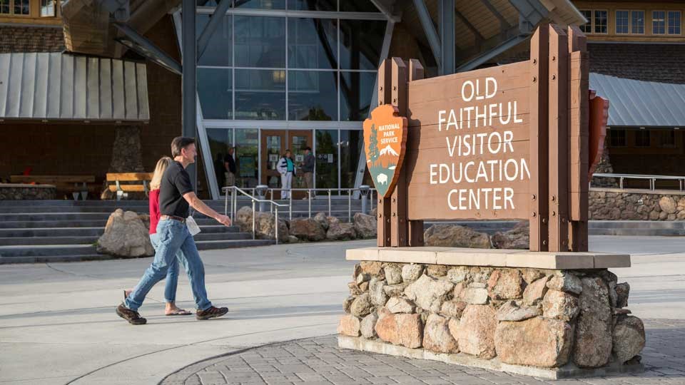 Two visitors walking by the sign in front of the wood, glass, and steel visitor center