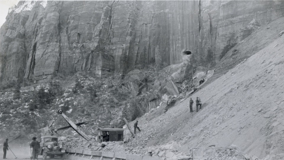 Black and white photo of men and machinery clearing a slope with a cliff in the background. A tunnel window is in the cliff.