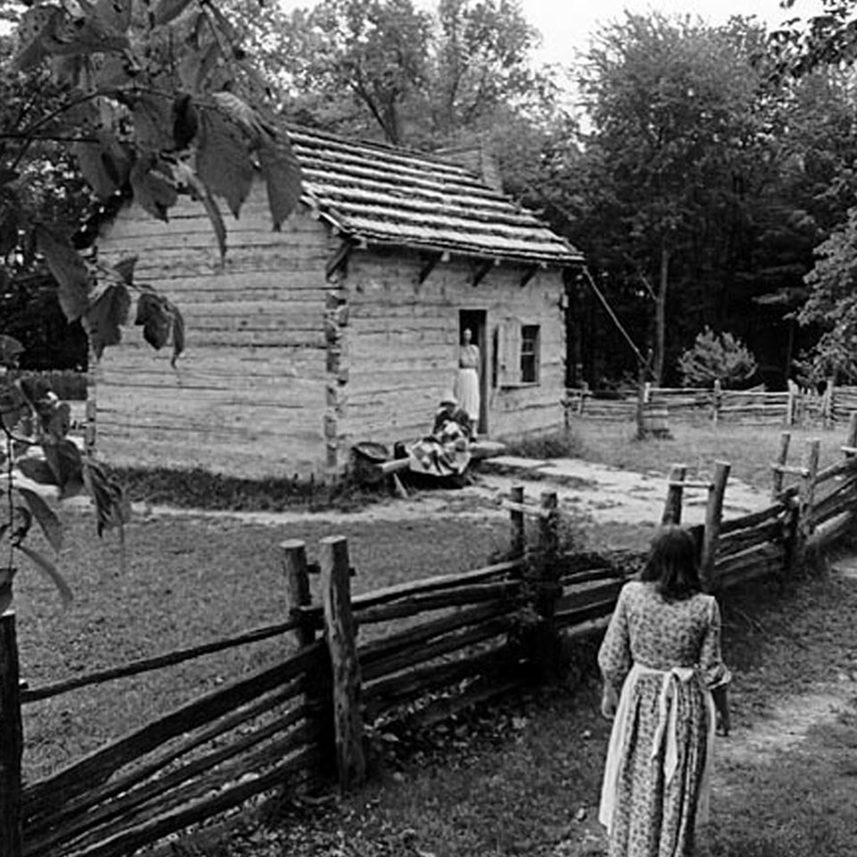 Black and white picture of a log cabin. One woman walking the trail to the cabin, the other at the doorway of the cabin. Split-rail fence in front.