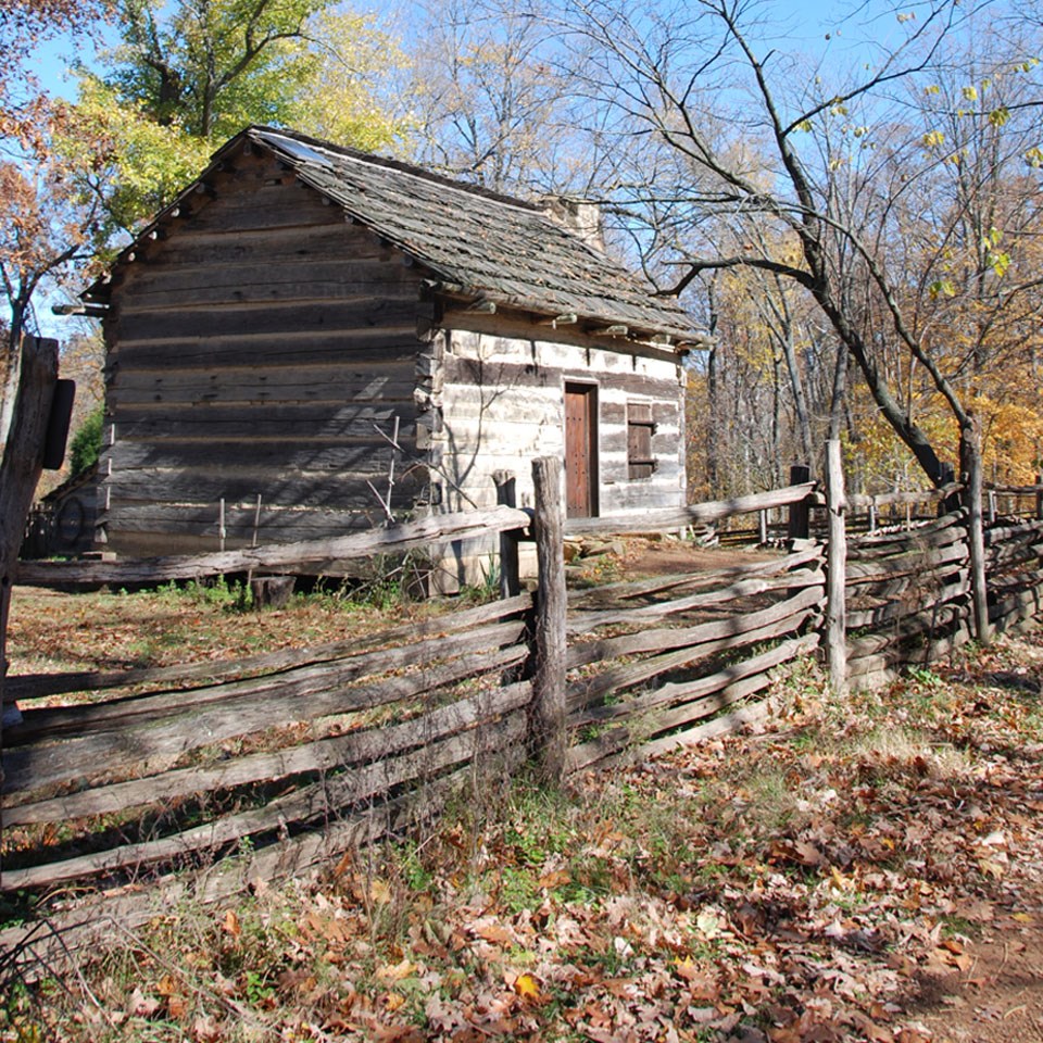 Black and white picture of a log cabin. One woman walking the trail to the cabin, the other at the doorway of the cabin. Split-rail fence in front.