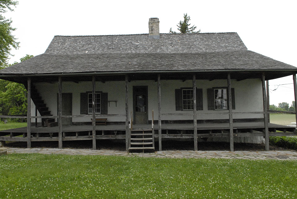 Black and white photo of a house with a front porch across its entire length and a central set of stairs.