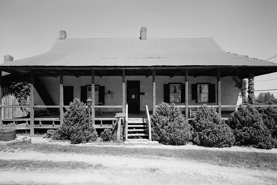 Black and white photo of a house with a front porch across its entire length and a central set of stairs.