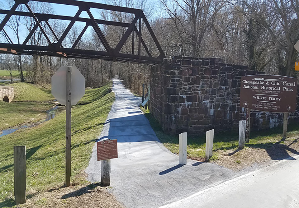 A photo of a dirt path surrounded by grass running beneath a metal bridge.