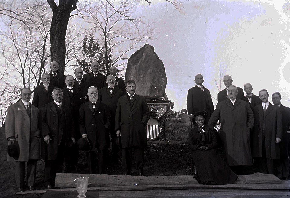 A group of men in dress coats and a seated woman in front of a stone marker