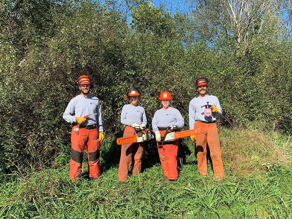 Two men and two women wearing hardhats and chaps and holding landscaping tools