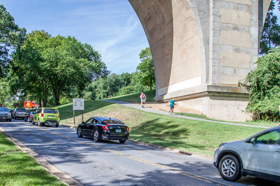 Road with potholes under bridge with footpath nearby.