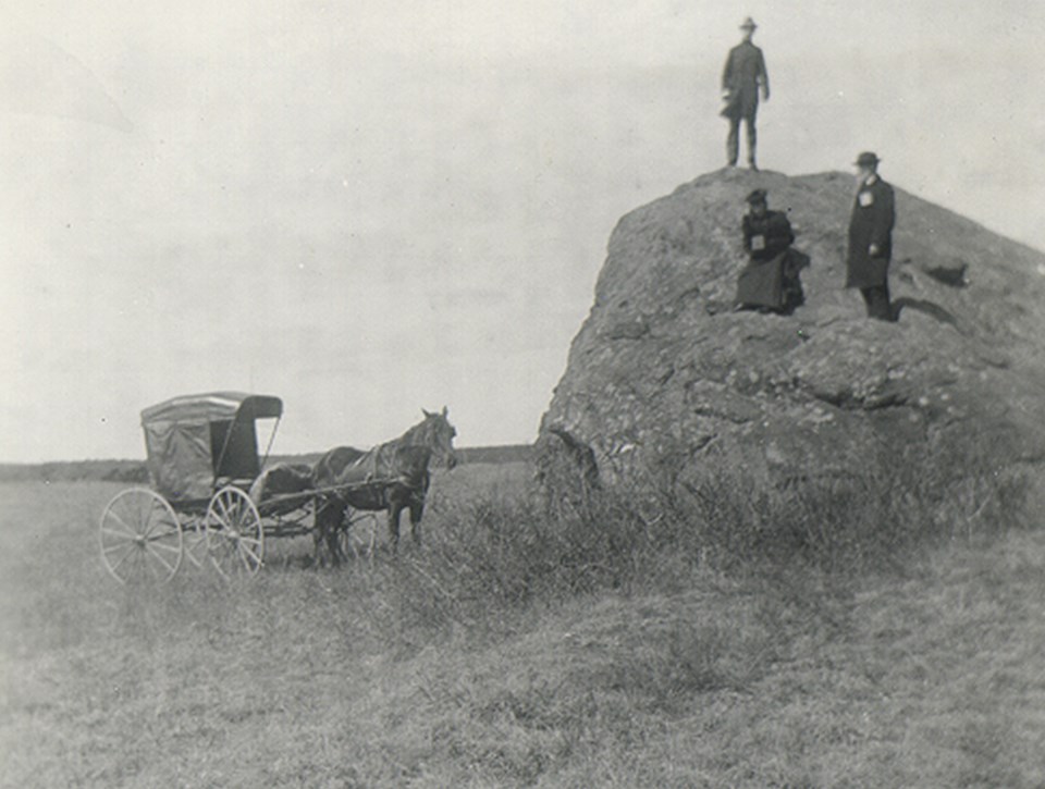 A black and white photo of three people sitting on a large boulder in an open field.