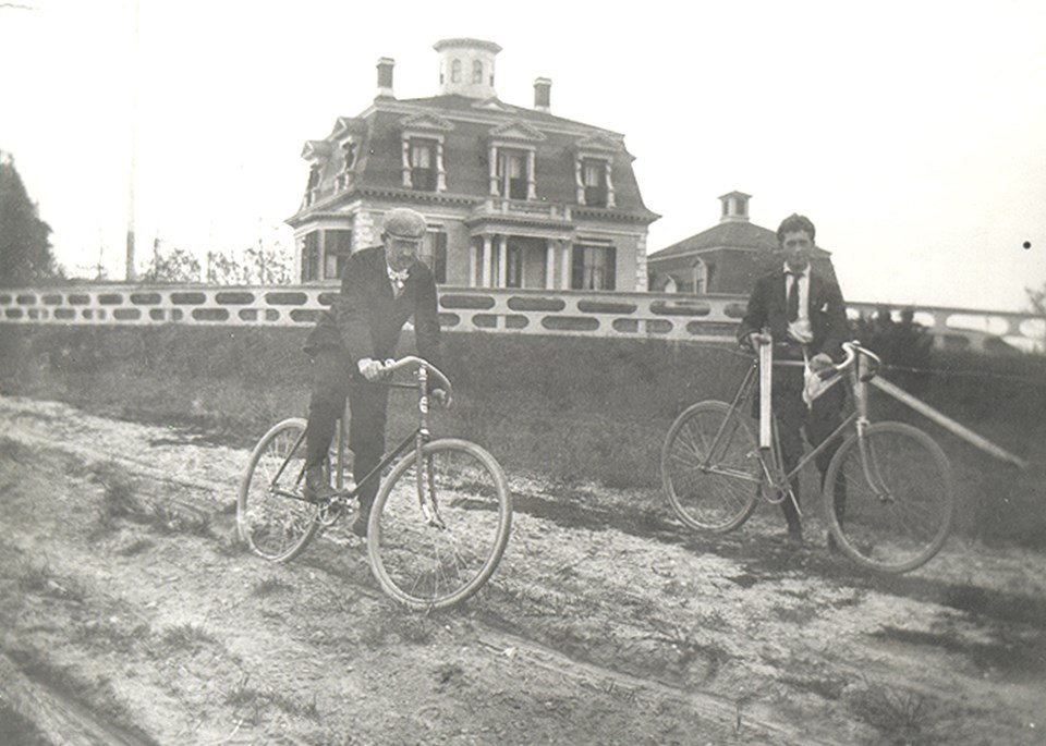 A man and a woman sit on bicycles in front of a bright yellow house on a hill.