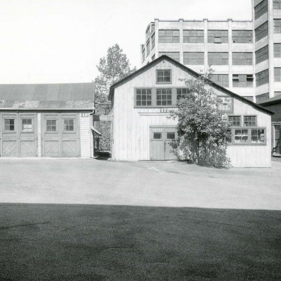 Black and white image of two wooden buildings. Building on left is rectangular, building on right is angular. Gravel and shadow in foreground. Large multi-story rectangular building in background.