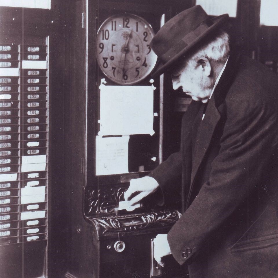 Black and white photo of Thomas Edison punching in at time clock, time cards to the left