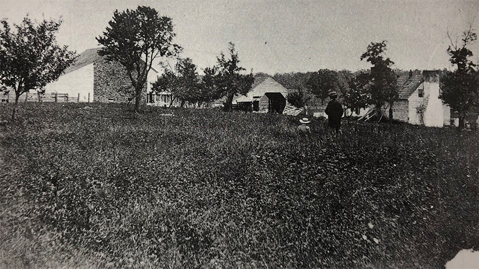 Two men look over a field at a large stone barn, a wagon shed, and a house.