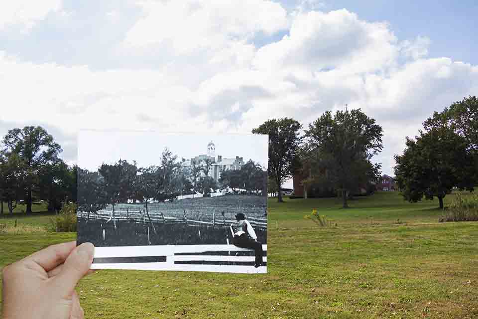 A young man sits on a white fence looking up the hill at a large building with a white cupola.
