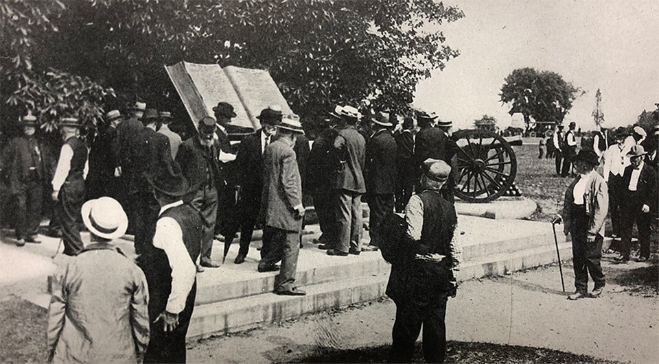 Civil War veterans gather around a large book shaped monument next to a grove of trees.