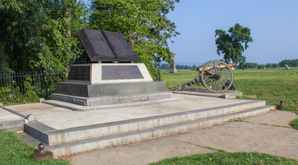 Civil War veterans gather around a large book shaped monument next to a grove of trees.