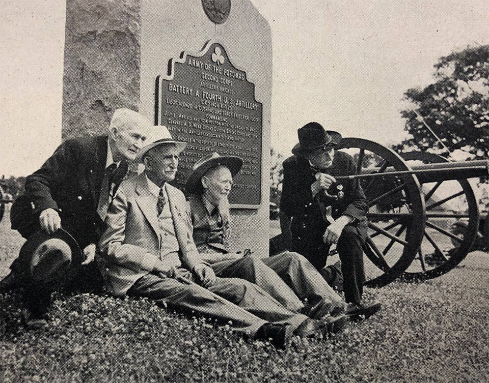 Four Civil War veterans sit at the base of a small monument as one of them points with his cane. A cannon sits in the background.