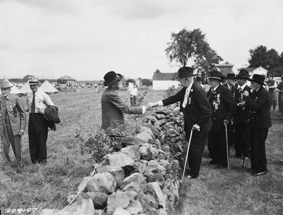 A Confederate veteran and a Union veteran shake hands over a stone wall. Other veterans watch on.