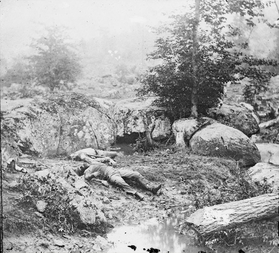 Two dead Confederate soldiers lie on the bank of a small pond, surrounded by large boulders.