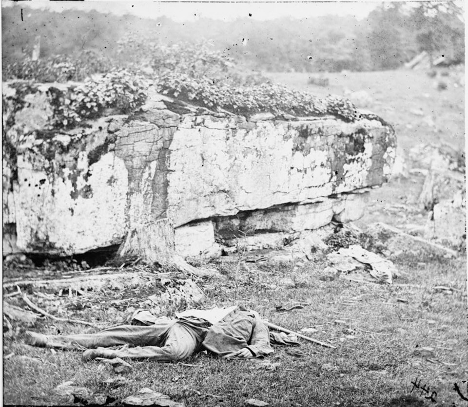 A black and white image of a dead man laying in front of a large boulder.