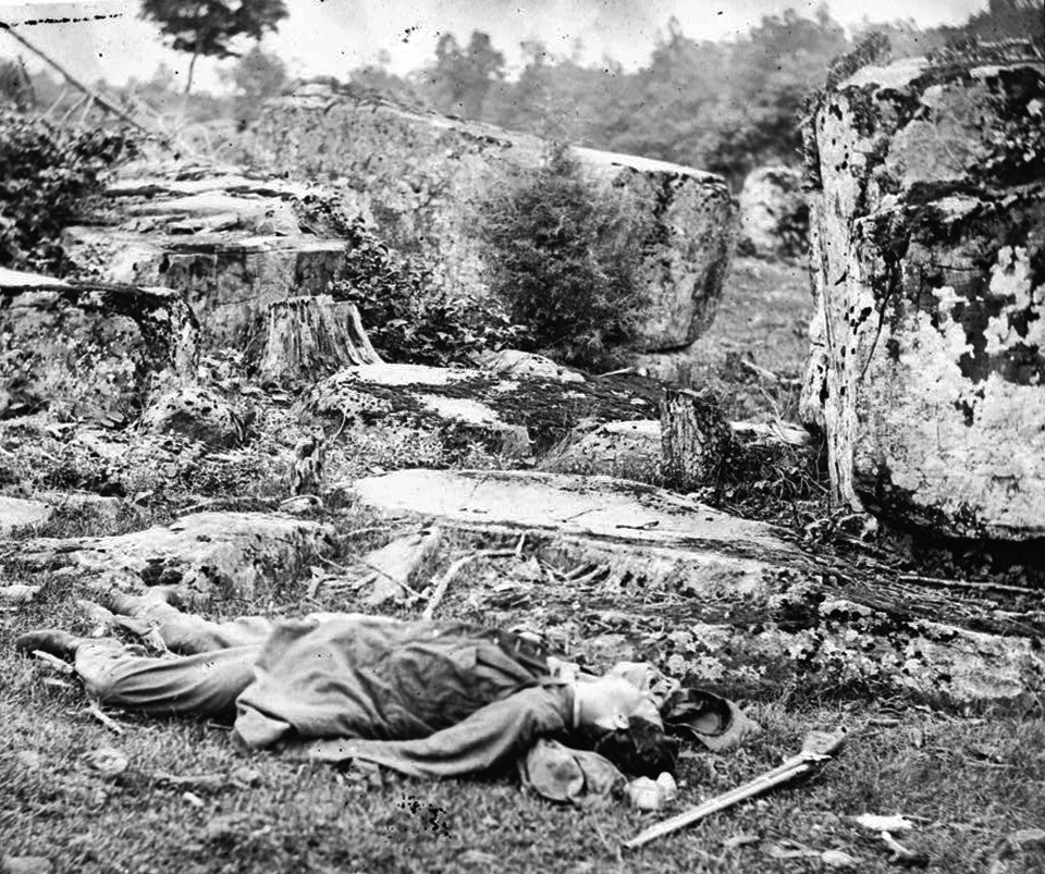 A dead Confederate soldier lies amidst debris near Devil’s Den, with a gun and hat near his head.
