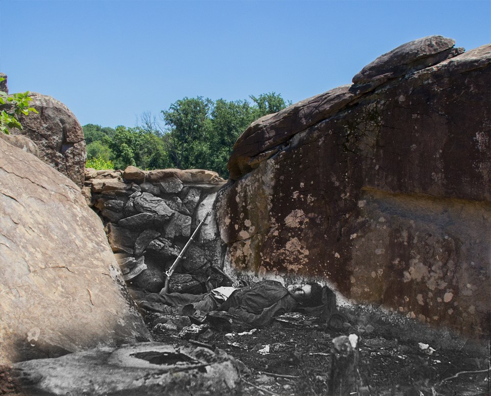 A dead Confederate soldier lies behind a stone fortification, a gun propped against the rocks next to him.