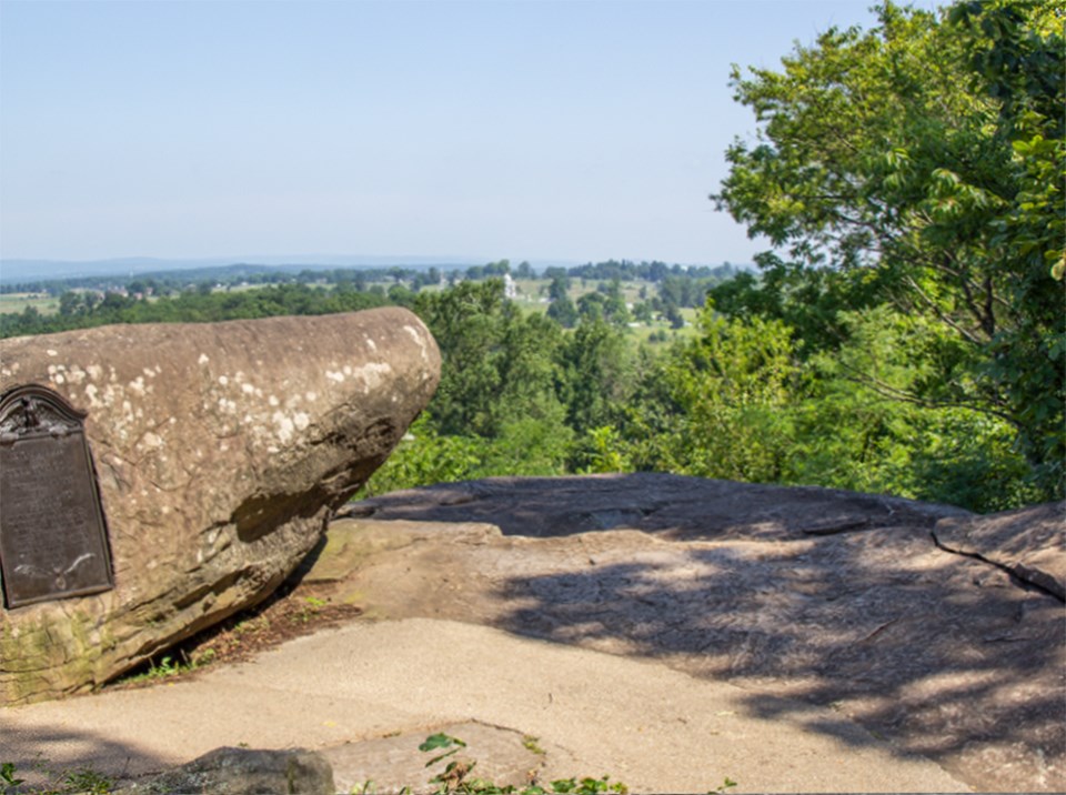 A large boulder is on the left and a pine tree is on the right in this black and white photo taken from the summit of Little Round Top.