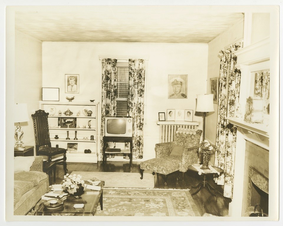 Floral curtains hang over two windows in the back of the room.  A TV set sits between the windows.  A gas fireplace is to the right.  A portrait of JFK is on the left back wall.  A chair and couch is to the left.