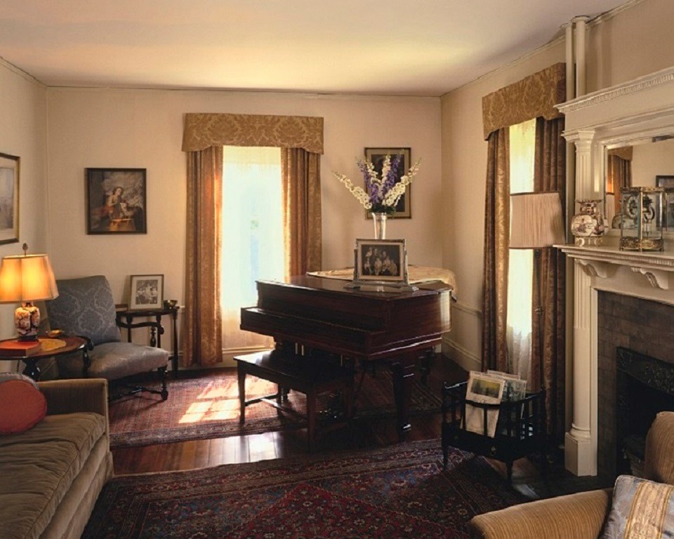 Floral curtains hang over two windows in the back of the room.  A TV set sits between the windows.  A gas fireplace is to the right.  A portrait of JFK is on the left back wall.  A chair and couch is to the left.