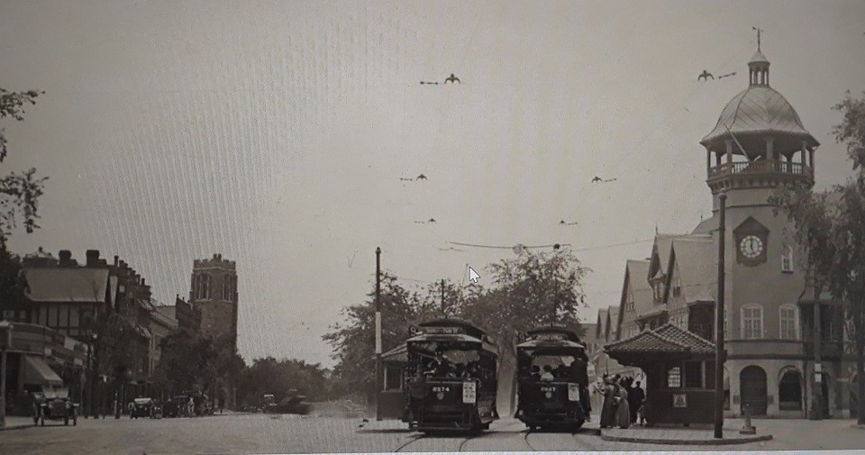 A view of Coolidge Corner at the intersection of Harvard and Beacon Streets.  The SS Pierce Building with clock tower is on the left.  A streetcar system runs alongside the SS Pierce to the left.  People are boarding the streetcar.