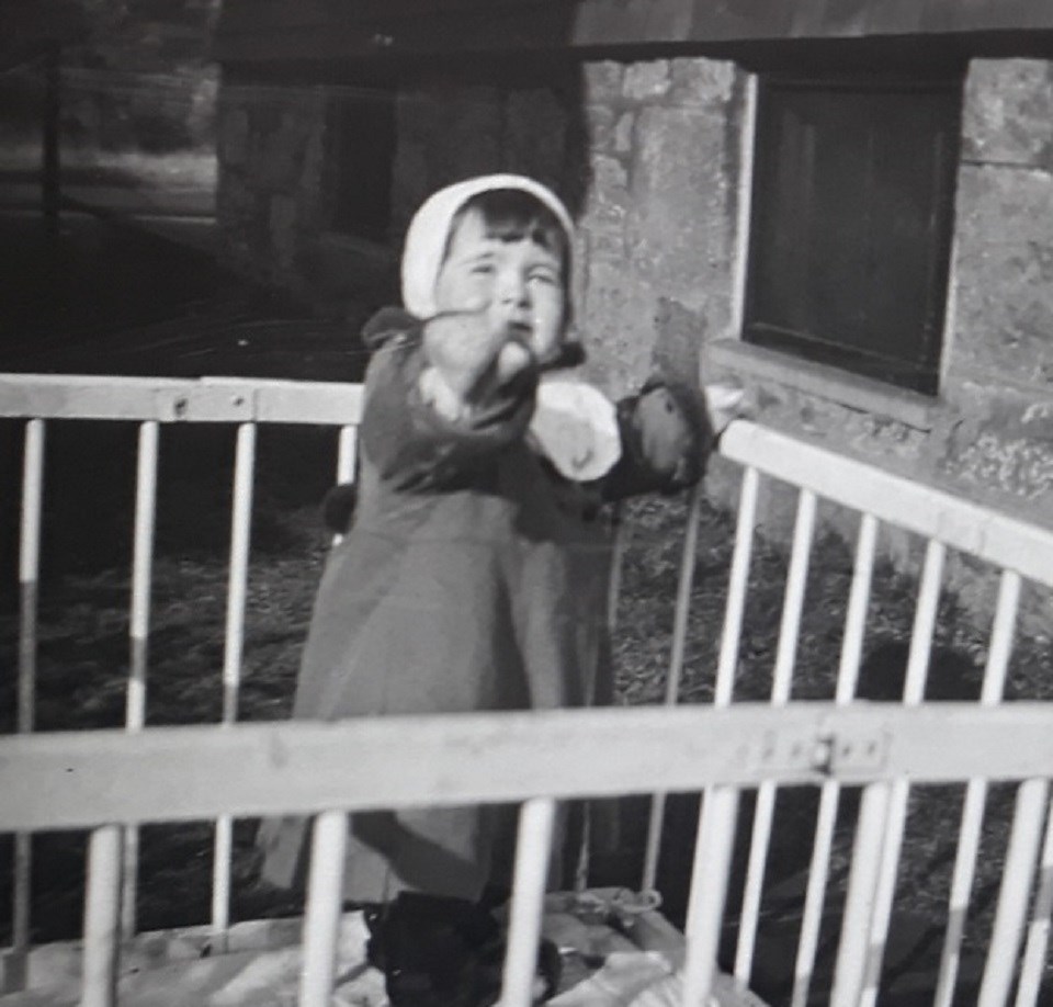 A black and white image of a toddler standing in a white playpen.  The play pen is near a stone foundation with windows.