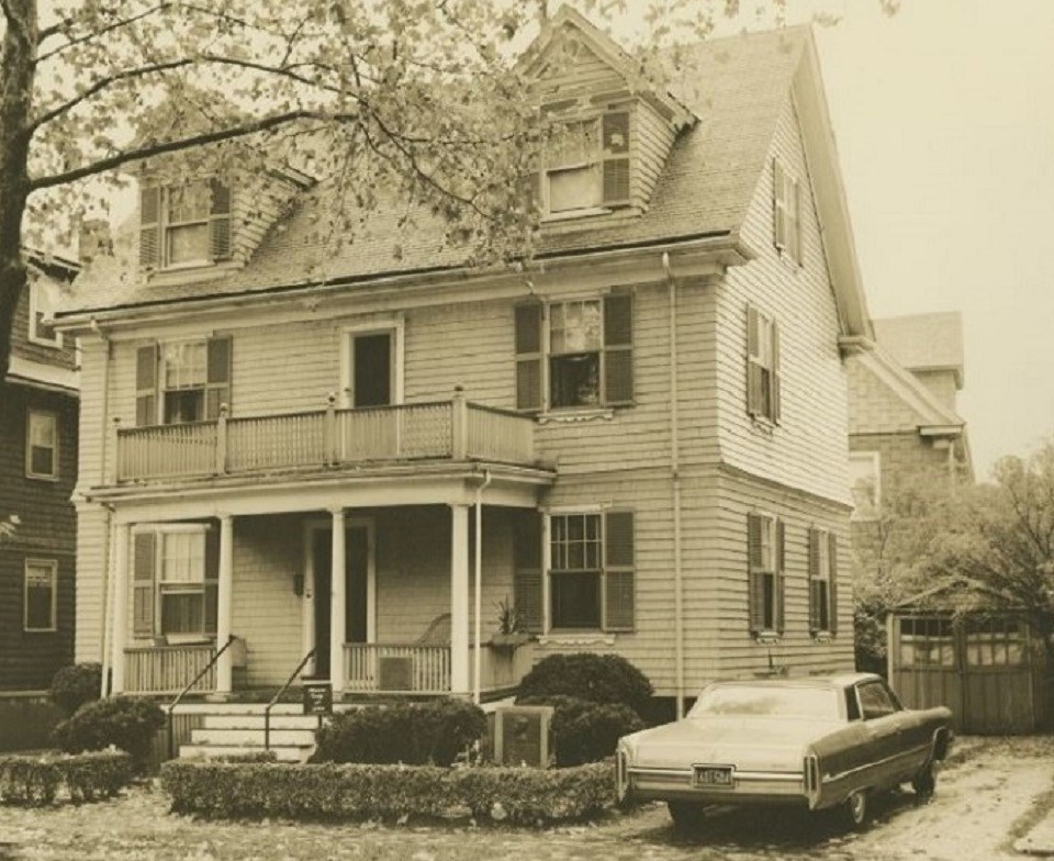 A black and white photo of the JFK Birthplace.  A light colored colonial house with dark shutters.  A tall leafless plane tree stands to the left front of the house.  A car sits on the right of the house.