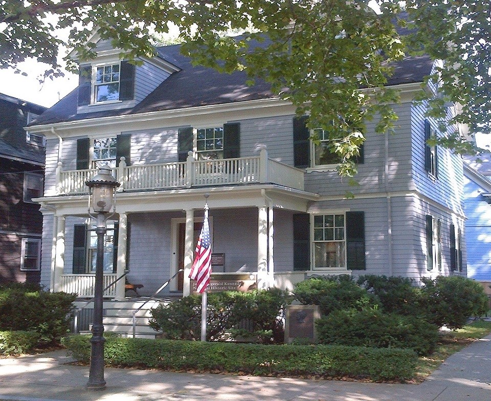 A black and white photo of the JFK Birthplace.  A light colored colonial house with dark shutters.  A tall leafless plane tree stands to the left front of the house.  A car sits on the right of the house.