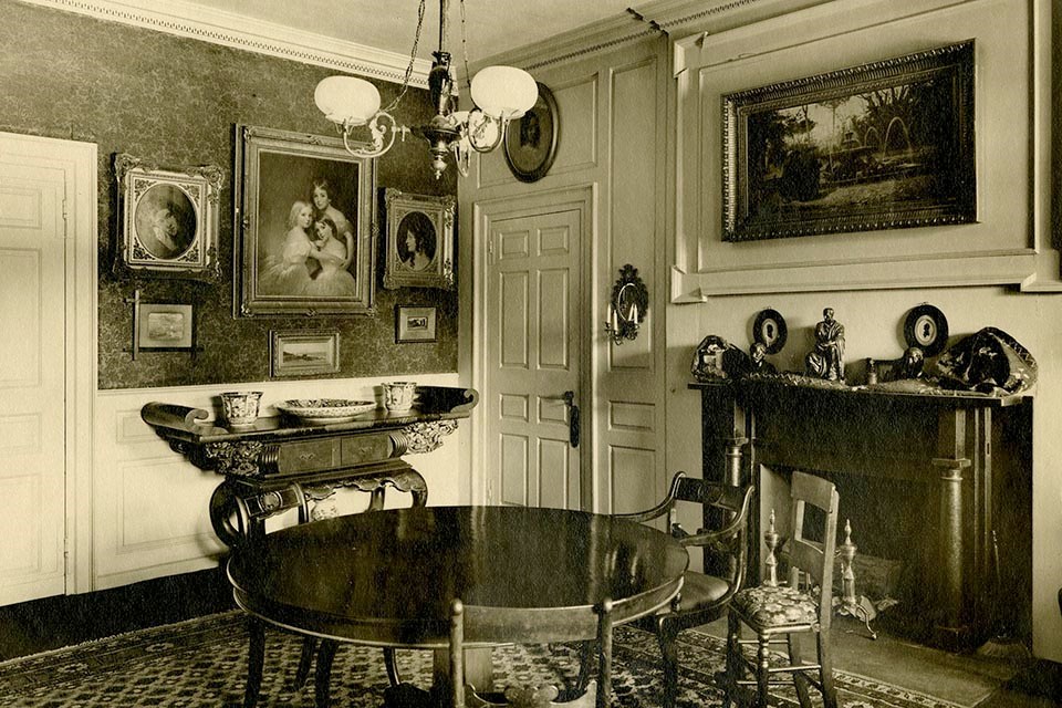 Black and white photograph of room with round table at center, paintings on walls